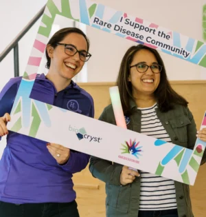 Two people smiling while holding a frame sign that reads, "I support the Rare Disease Community," with logos for BioCryst and rarediseaseday.org.