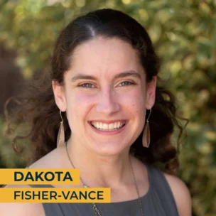 A woman with curly brown hair, wearing a gray dress and long earrings, smiles at the camera. Text on the image reads: "Dakota Fisher-Vance".