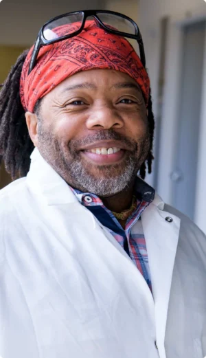 A man wearing a red bandana, glasses on his head, and a white lab coat, smiles while standing indoors.