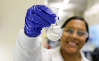 A scientist wearing a white lab coat and safety glasses holds up a round-bottom flask containing a white substance, with a blue gloved hand.