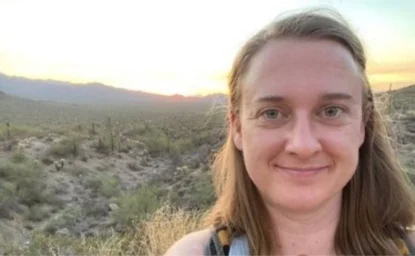 A woman with long hair is smiling for a selfie in a desert landscape at sunset. Cacti and dry vegetation are visible in the background.