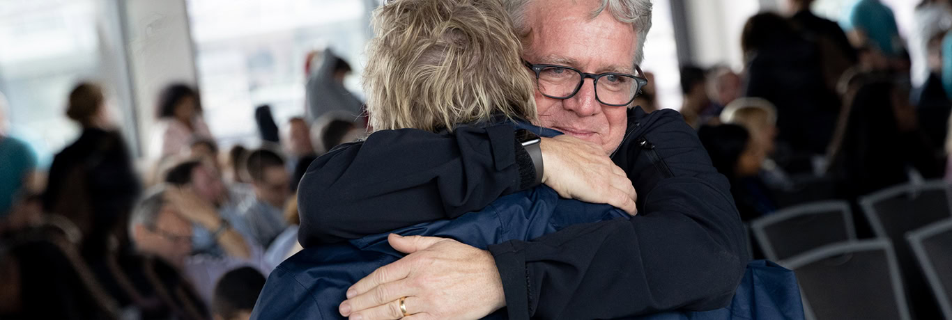 Two people embrace tightly in a hug at a crowded indoor event, surrounded by several others in the background.