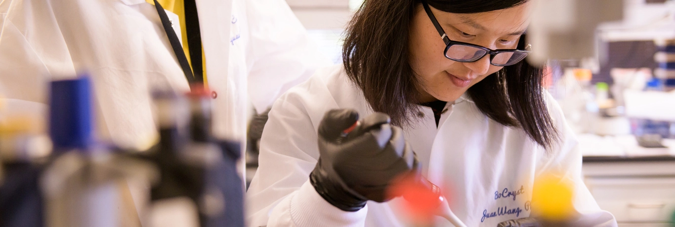 A person in a lab coat and gloves uses a pipette at a laboratory workstation, with equipment visible in the background.