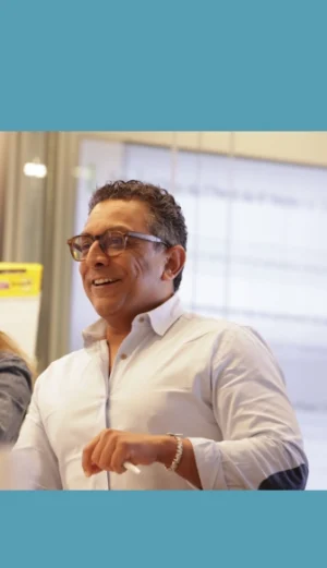 A man wearing glasses and a white shirt is smiling and standing indoors, with part of a screen visible in the background.