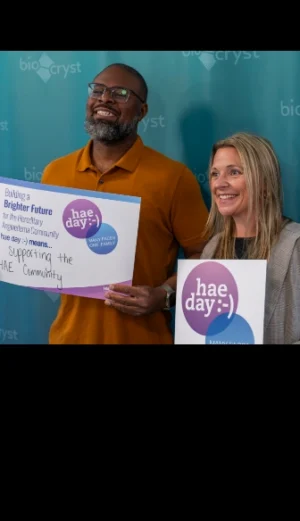Two people stand in front of a blue backdrop holding signs that promote support for the HAE community and awareness for hae day.
