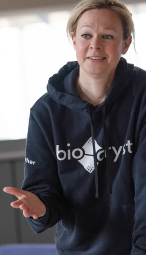 A person wearing a navy "bio-cryst" hoodie gestures with one hand while speaking indoors.