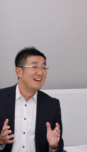 A man in a suit and glasses sits indoors, smiling and gesturing with his hands while talking.