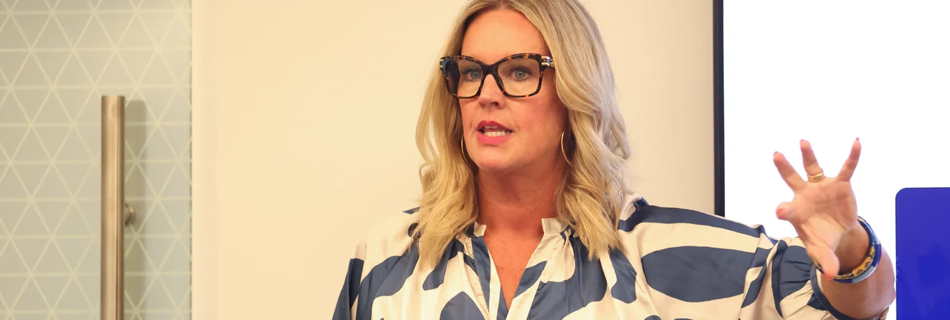 A woman with blonde hair and glasses speaks while gesturing with her hand. She is wearing a blue and white patterned shirt and stands in front of a light-colored wall.