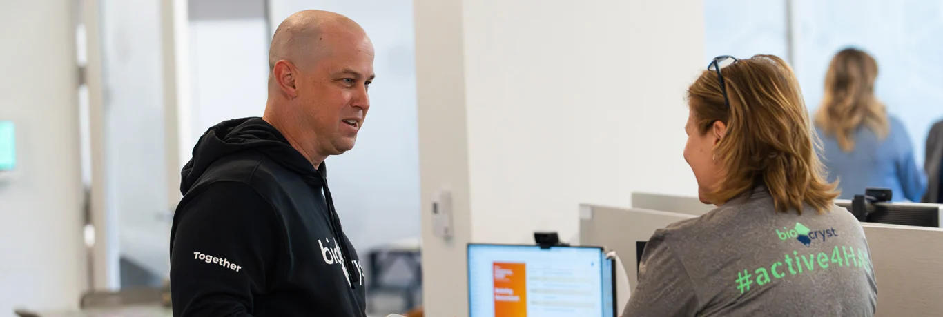 Two people are having a conversation in an office setting; one is standing and wearing a black hoodie, while the other sits at a desk wearing a gray t-shirt with text on the back.