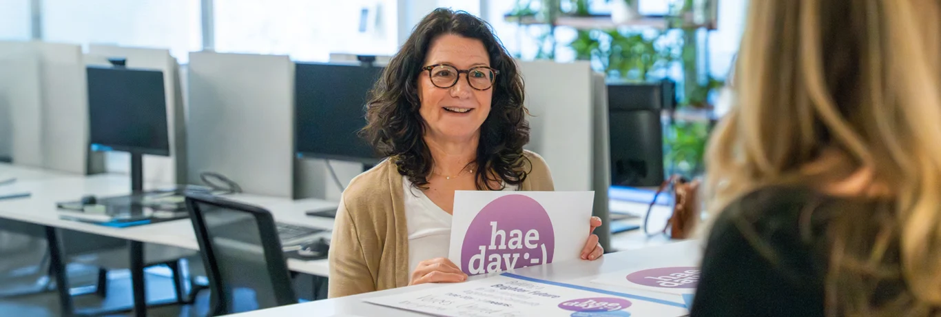 A woman sits at a desk in an office holding a sign that reads “hae day :-)” while talking to another person.