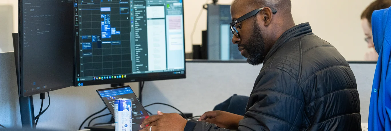 A man in a black jacket works at a desk with a laptop and two large monitors displaying technical data in an office setting.