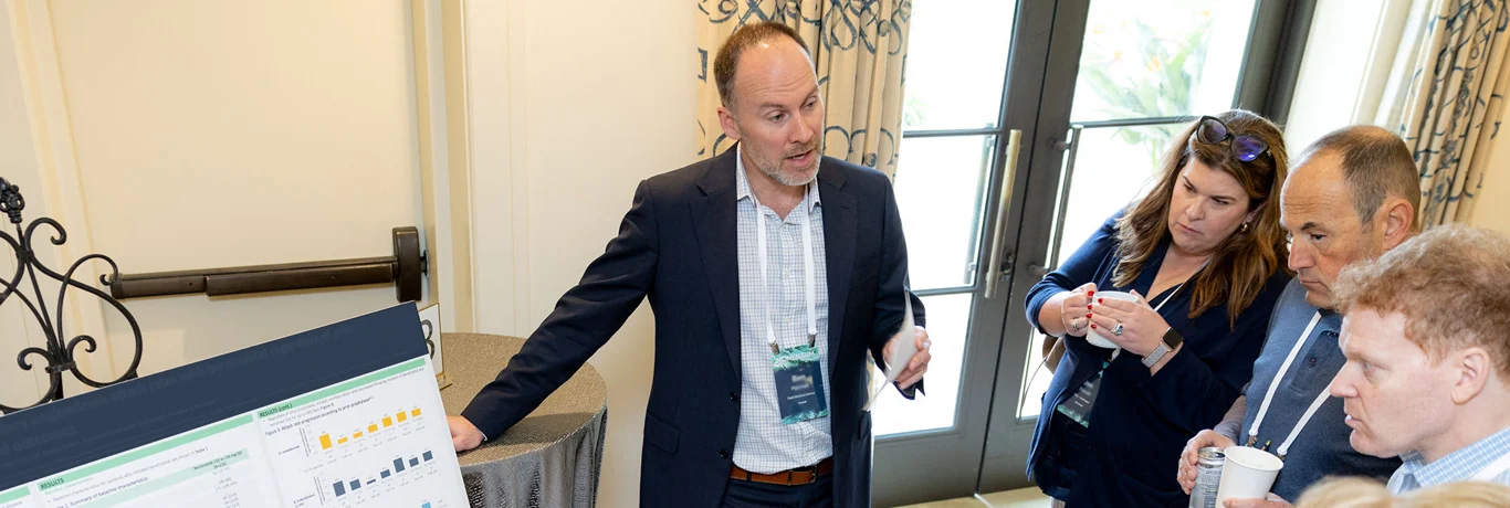 Man in a suit presents data on a poster board to three attentive people in a conference room, two holding cups and one taking notes.