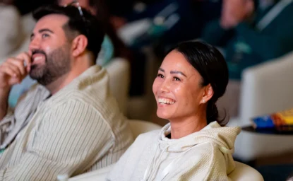 Two people sit side by side, smiling and looking forward, appearing engaged, in what seems to be an indoor event or presentation.