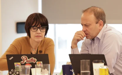 Two people sit at a table working on laptops, focused on their screens. Various drinks and office supplies are visible on the table.
