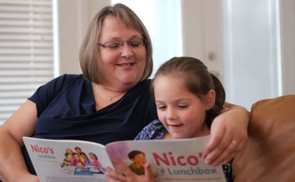 An adult woman and a young girl sit on a couch reading a book titled "Nico's Lunchbox" together, both smiling.