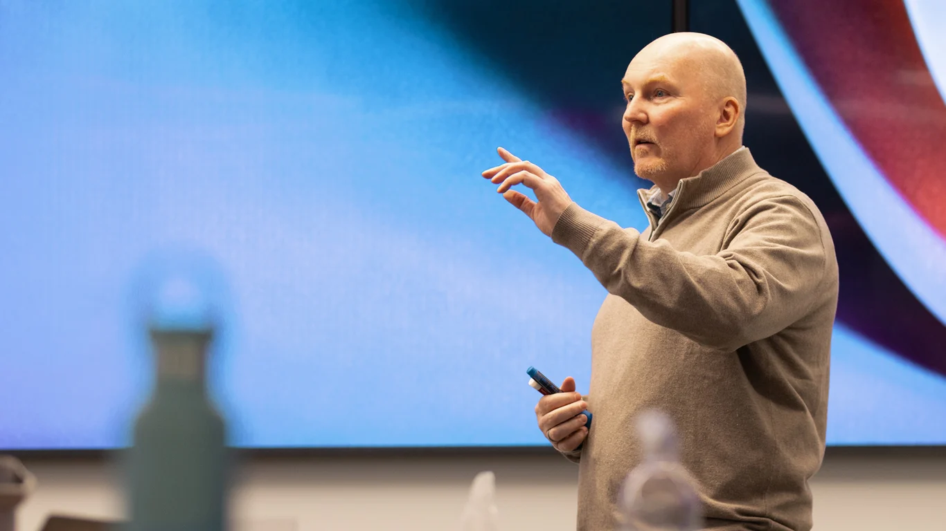 A man in a beige sweater speaks and gestures while holding a marker in front of a large screen during a presentation.