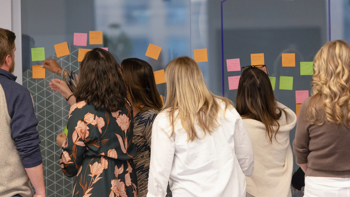 Six people stand facing a glass wall covered with colorful sticky notes, appearing to collaborate or brainstorm in an office setting.