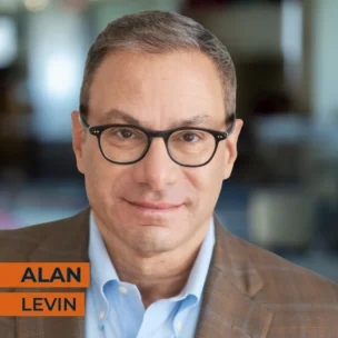 Man with short brown hair and glasses wearing a plaid blazer and blue shirt, standing indoors. Text overlay reads "ALAN LEVIN.