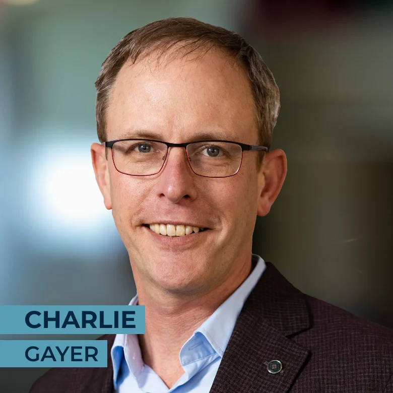 A man wearing glasses and a suit jacket smiles at the camera. Text on the image reads "Charlie Gayer.