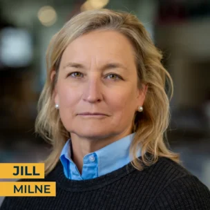 A woman with blonde hair, wearing a blue collared shirt and dark sweater, looks at the camera. Text in the image reads "Jill Milne.