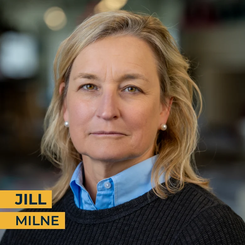 A woman with blonde hair, wearing a blue collared shirt and dark sweater, looks at the camera. Text in the image reads "Jill Milne.
