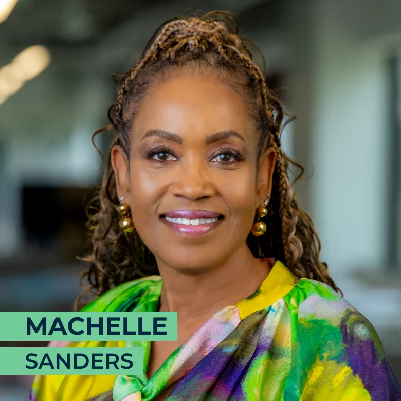 A woman with braided hair and gold earrings wearing a colorful blouse, smiling at the camera in an indoor office setting. The text "Machelle Sanders" appears on the image.