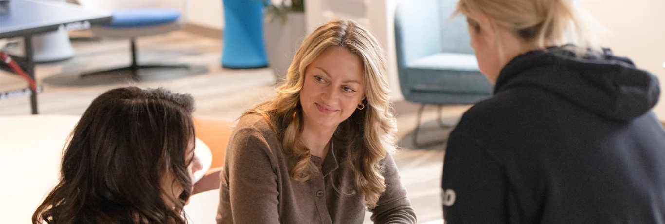 Three women sit together at a table in an office setting, engaged in conversation.