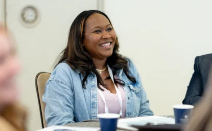A woman with long dark hair wearing a denim jacket smiles while seated at a table during a meeting.