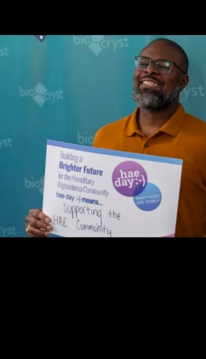 A man holding a sign about HAE Day, standing in front of a blue background with “bioCryst” logos, smiles at the camera.